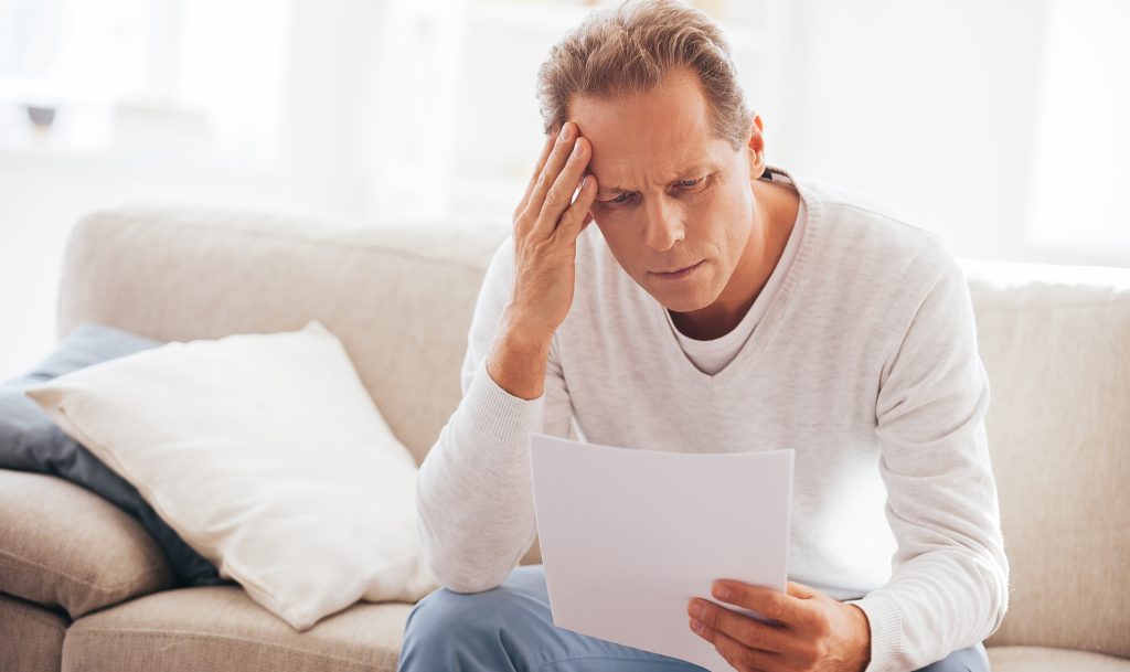 Depressed mature man holding paper and looking at it while sitting on the couch at home