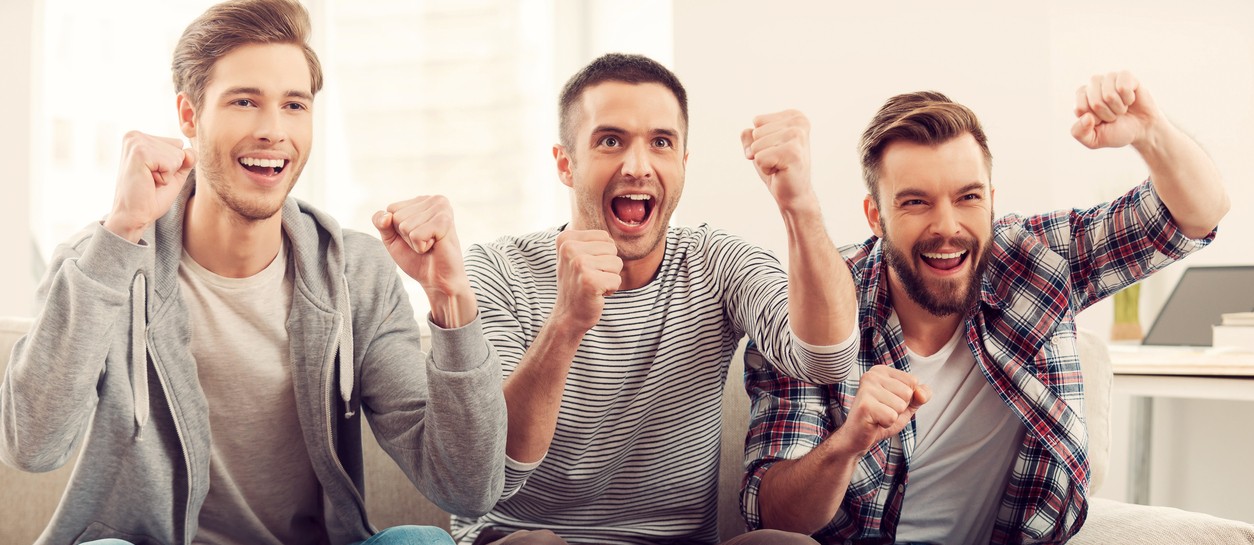 Three happy young men watching football game and keeping arms raised while sitting on sofa